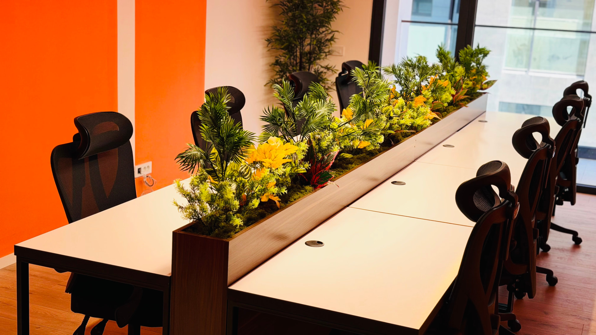 Long coworking desk with orange wall, plants, and office chairs at PRN CO-WORK